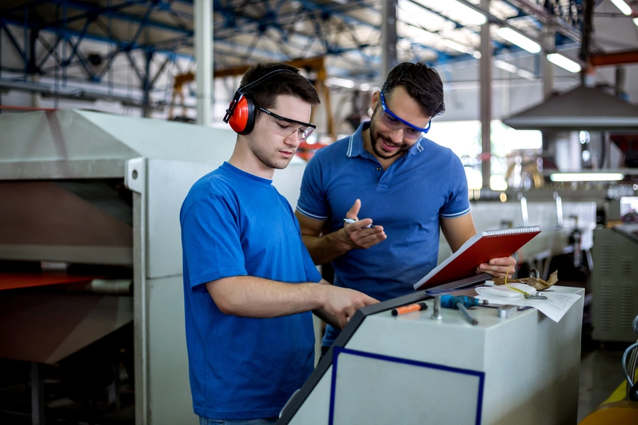 zarządzanie jakością (dwóch mężczyzn w niebieskich koszulkach przy maszynie, jeden ma słuchawki tłumiące)