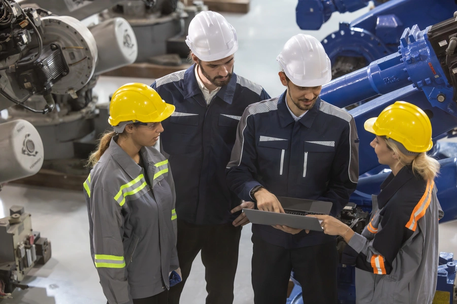 transformacja cyfrowa: dwóch mężczyzn w białych kaskach i dwie kobietry w żółtych kaskach stoją na hali produkcyjnej i analizują dane z laptopa